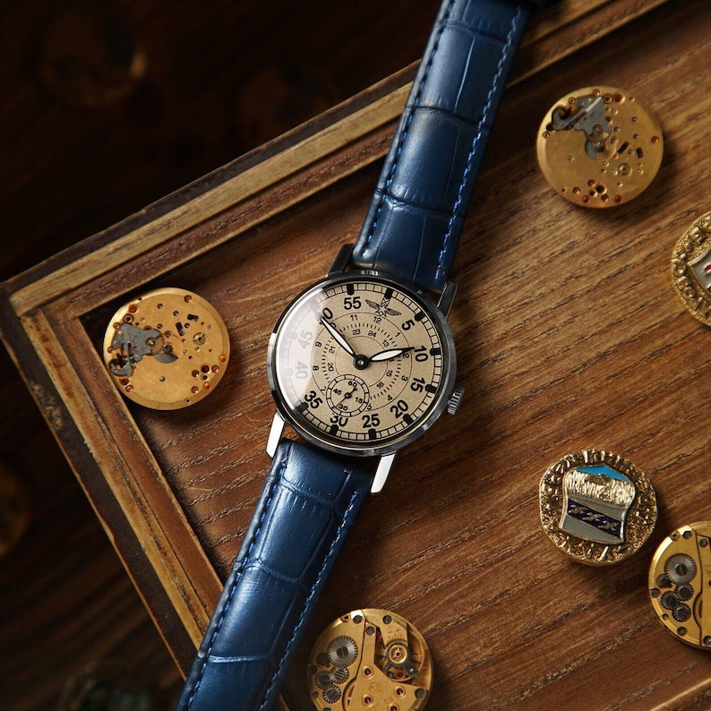 May include: A silver wristwatch with a blue leather band and a cream-colored face. The watch face has black Arabic numerals and a black second hand. The watch is on a wooden surface with other watch parts.