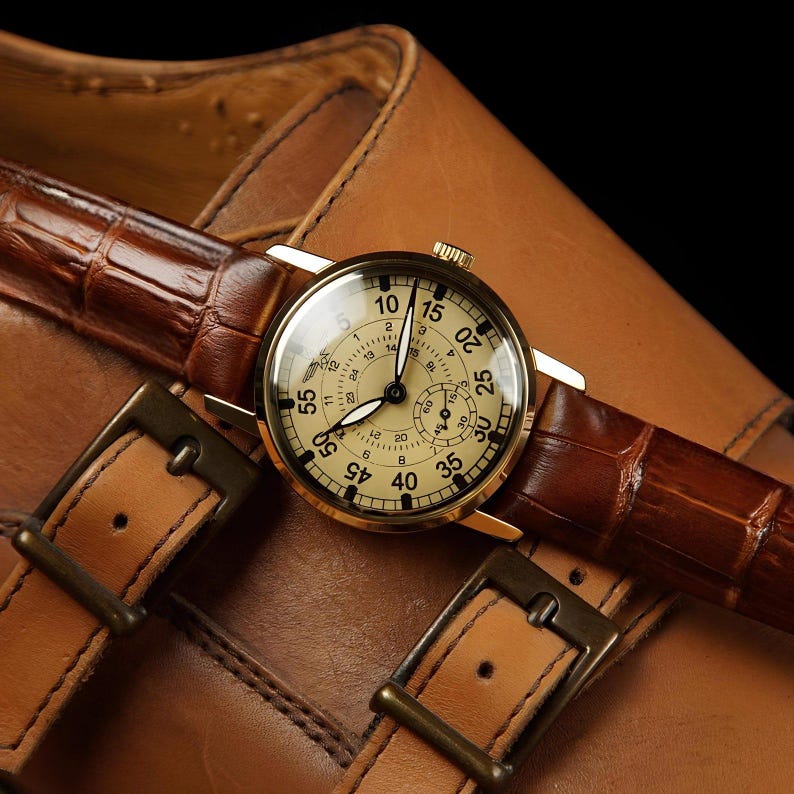 May include: A gold-tone wristwatch with a cream-coloured dial featuring black numerals, a sub-dial, and a brown alligator-textured leather strap. The watch face displays a 12-hour and 24-hour scale with a small winged logo near the top centre.