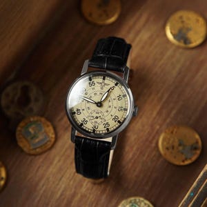 Puede incluir: Un reloj de pulsera de estilo vintage con esfera color crema, correa de cuero negro y caja plateada. El reloj presenta m&uacute;ltiples esferas y n&uacute;meros, incluido un peque&ntilde;o dial de segundos. El reloj se exhibe sobre una superficie de madera con monedas doradas.