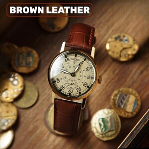 Peut inclure: Une montre dor&eacute;e avec un cadran cr&egrave;me et un bracelet en cuir marron. Le cadran de la montre affiche des chiffres et des sous-cadrans. Le texte "BROWN LEATHER" est en haut. La montre est entour&eacute;e de pi&egrave;ces de monnaie anciennes sur une surface en bois.