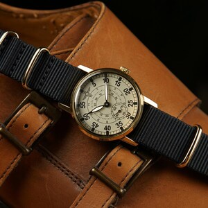 May include: A gold-toned wristwatch with a cream-colored face and black strap. The watch features black numerals and hands, with a smaller dial at the bottom. The watch is set against a brown leather background, suggesting a classic or vintage style.