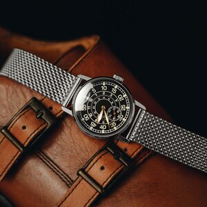 May include: A silver-toned wristwatch with a black face and a mesh band rests on a brown leather surface. The watch face displays white numerals and hands. The leather surface features two buckles.