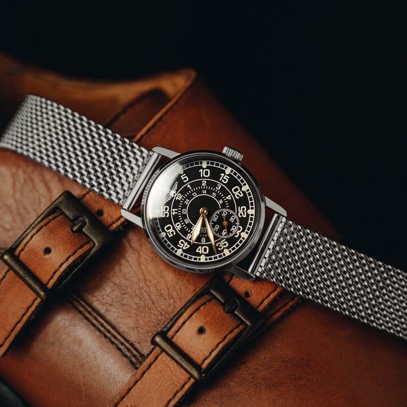 May include: A silver-toned wristwatch with a black face and a mesh band rests on a brown leather surface. The watch face displays white numerals and hands. The leather surface features two buckles.