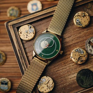 May include: A gold-toned wristwatch with a green face and a mesh band. The watch face features a moon phase design with a red second hand. The watch is displayed on a wooden surface with watch components and decorative coins.
