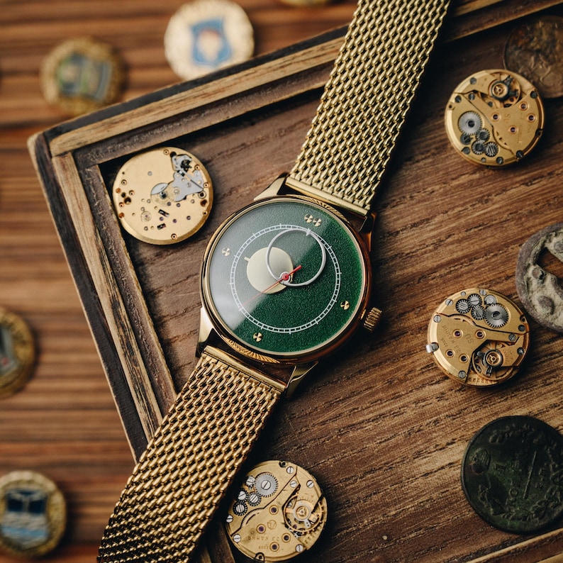 May include: A gold-toned wristwatch with a green face and a mesh band. The watch face features a moon phase design with a red second hand. The watch is displayed on a wooden surface with watch components and decorative coins.