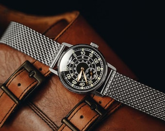 May include: A wristwatch with a round black dial featuring white Arabic numerals for minutes and a smaller inner circle for hours. The watch has gold-toned hands and a silver-toned stainless steel mesh strap with a polished finish and a circular case.