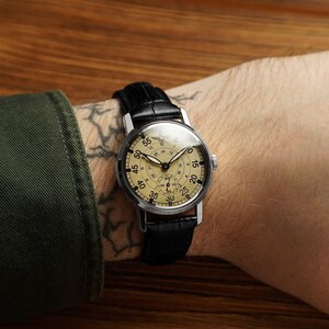 May include: A vintage-style wristwatch with a cream-colored face, black numerals, and a black leather band. The watch has a silver-colored case and a second dial. The watch is on a wrist.