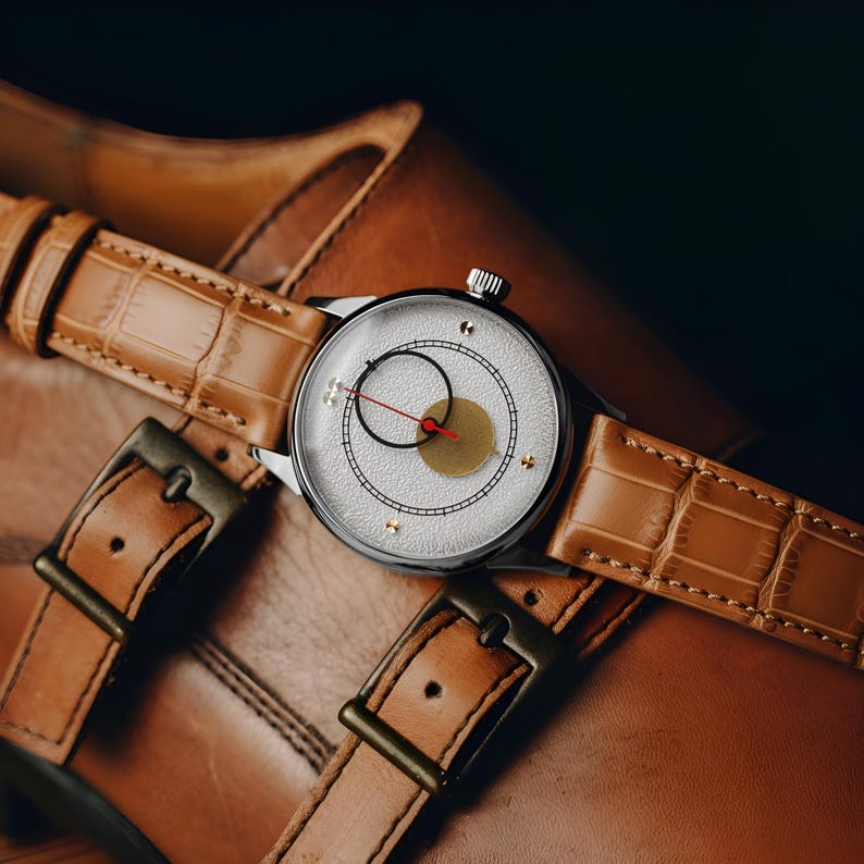 May include: A silver wristwatch with a white face, black and gold accents, and a red second hand rests on a brown leather strap. The watch is set on a pair of brown leather shoes with buckle closures, showcasing a classic style.