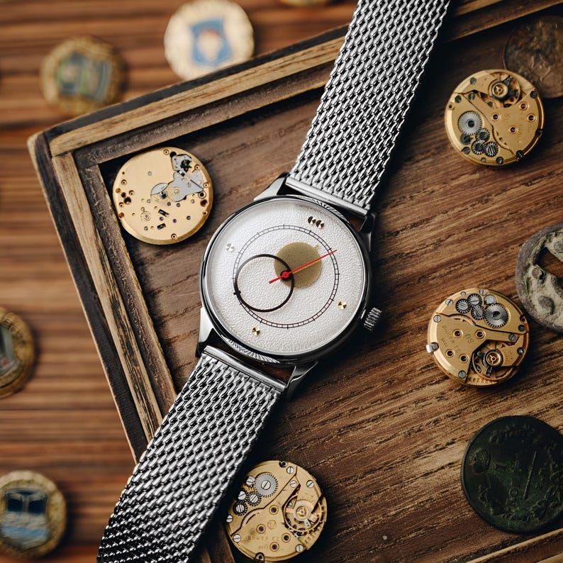 May include: A silver wristwatch with a mesh band and a white face featuring a black circular design and a red hand. The watch is displayed on a wooden surface with watch components and vintage coins.