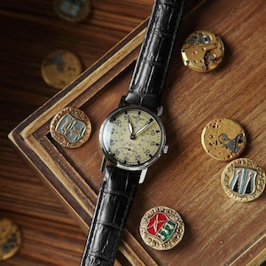 May include: A vintage-style wristwatch with a black leather band and a cream-colored face. The watch is surrounded by antique gold-colored coins and watch parts, set on a wooden surface. The watch face features black numerals and hands.