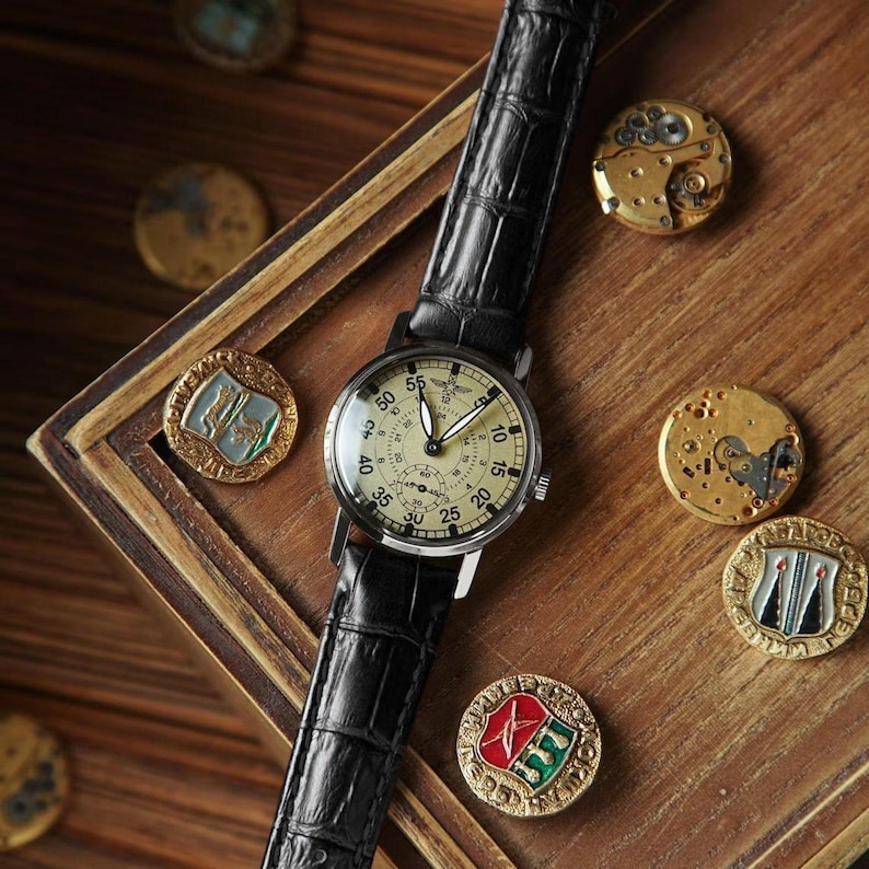 May include: A vintage-style wristwatch with a black leather band and a cream-colored face. The watch is surrounded by antique gold-colored coins and watch parts, set on a wooden surface. The watch face features black numerals and hands.