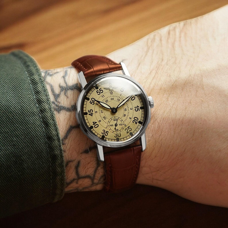 Puede incluir: Un reloj de pulsera de estilo vintage con esfera color crema y correa de cuero marr&oacute;n. El reloj tiene una caja de acero inoxidable y presenta m&uacute;ltiples subesferas y n&uacute;meros negros. La correa mide aproximadamente 2 cm de ancho.