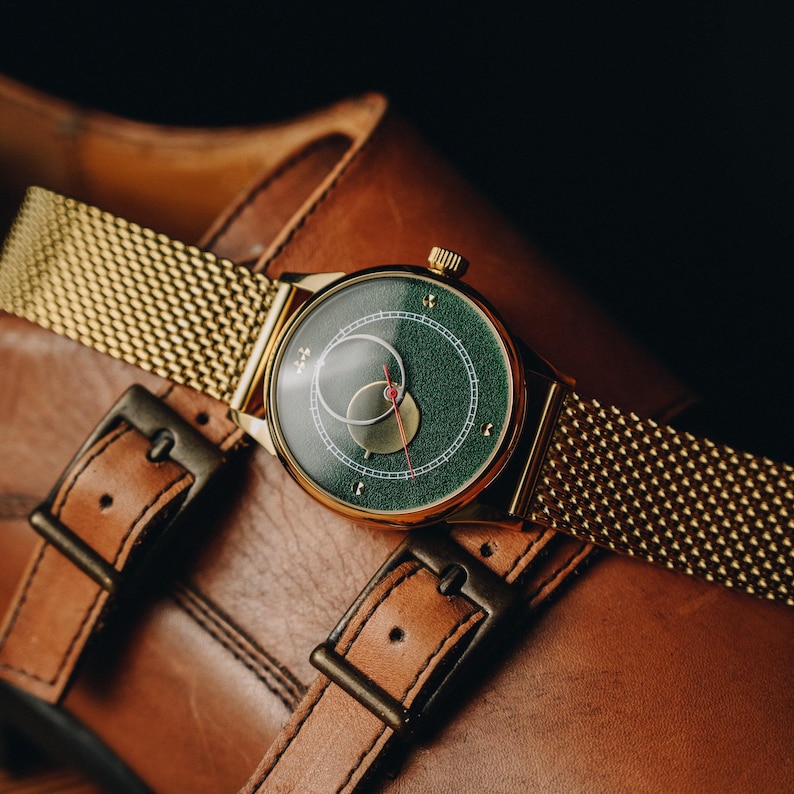 May include: A gold-toned wristwatch with a green face and a gold mesh band rests on a brown leather shoe. The watch face features white and gold accents with a red second hand. The shoe has two buckles.