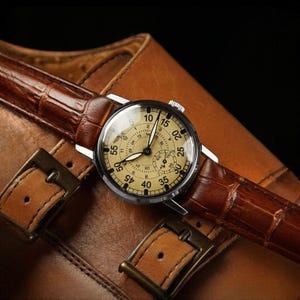Puede incluir: Un reloj de estilo vintage con esfera crema y n&uacute;meros negros, con una correa de cuero marr&oacute;n con estampado de cocodrilo. El reloj est&aacute; sobre una superficie de cuero marr&oacute;n, con hebillas met&aacute;licas visibles. El di&aacute;metro de la esfera es de aproximadamente 4 cm.