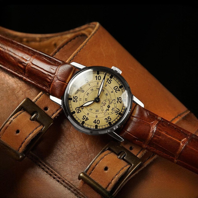 Puede incluir: Un reloj de estilo vintage con esfera crema y n&uacute;meros negros, con una correa de cuero marr&oacute;n con estampado de cocodrilo. El reloj est&aacute; sobre una superficie de cuero marr&oacute;n, con hebillas met&aacute;licas visibles. El di&aacute;metro de la esfera es de aproximadamente 4 cm.
