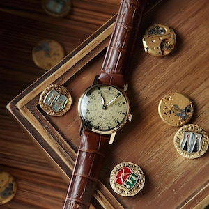 K&ouml;nnte beinhalten: Eine Vintage-Armbanduhr mit cremefarbenem Zifferblatt und braunem Lederarmband. Die Uhr liegt auf einer Holzoberfl&auml;che, umgeben von dekorativen goldfarbenen Medaillons und Uhrwerksteilen. Das Zifferblatt zeigt Zahlen und Hilfszifferbl&auml;tter.