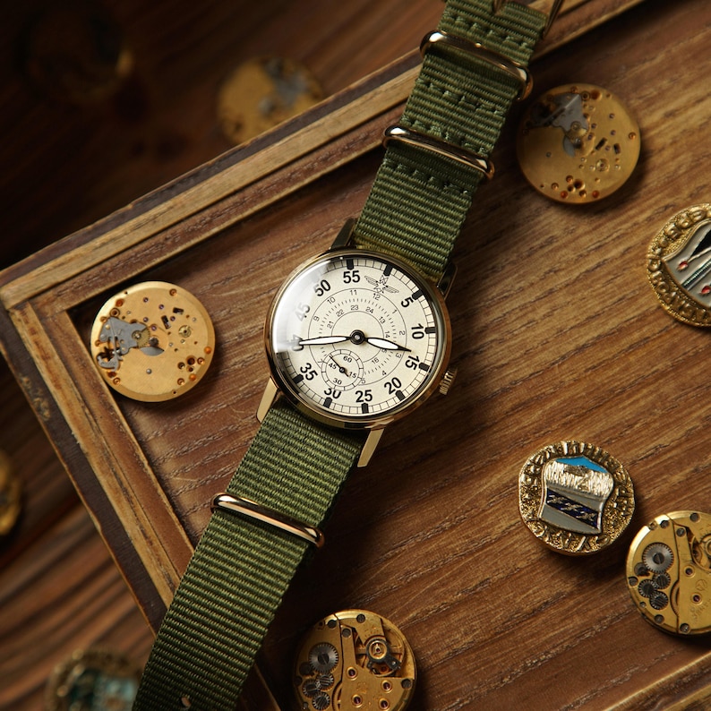 May include: A vintage-style wristwatch with a cream-colored face and black numerals, set in a gold-toned case. It features a green nylon strap and is displayed on a wooden surface with watch parts and decorative pins.
