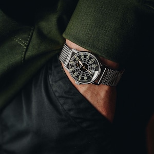 May include: A close-up of a wristwatch with a black face and silver mesh band. The watch has white numerals and hands, with a second hand. The watch is on a wrist, with a green long-sleeve shirt and black pants in the background.