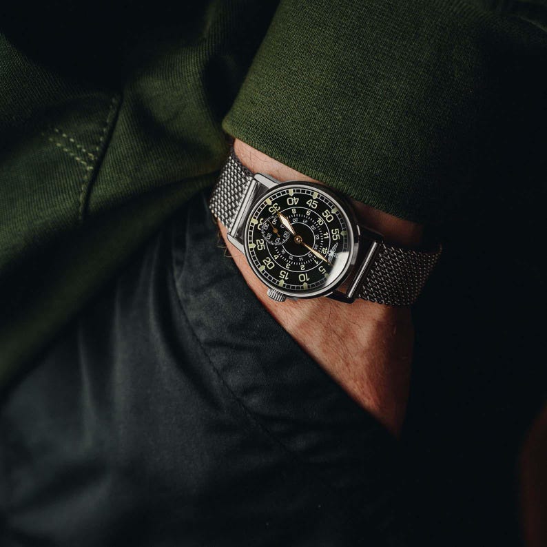May include: A close-up of a wristwatch with a black face and silver mesh band. The watch has white numerals and hands, with a second hand. The watch is on a wrist, with a green long-sleeve shirt and black pants in the background.