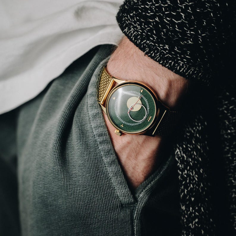 May include: A gold-toned wristwatch with a green face and a mesh band. The watch face features a circular design with a light-colored center and a small red accent. The watch is worn on a wrist, with a portion of a sleeve and sweater visible.
