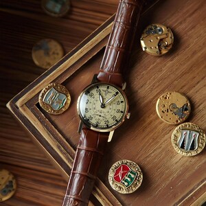 May include: A wristwatch with a cream-coloured dial, black numerals, and a brown leather strap with a crocodile texture. The watch features a central winged emblem and a sub-dial. Several circular metal watch components and pins are arranged nearby.