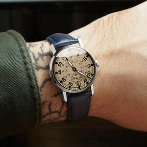 May include: A silver wristwatch with a cream-colored face and black markings. The watch has a blue leather band and a 24-hour dial.