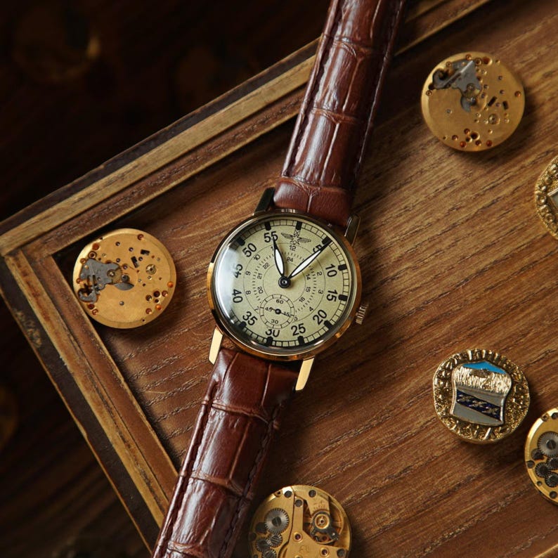 May include: A classic wristwatch featuring a brown leather strap and a gold-toned case. The watch face is cream with black numerals and hands. The watch is displayed on a wooden surface with watch components.