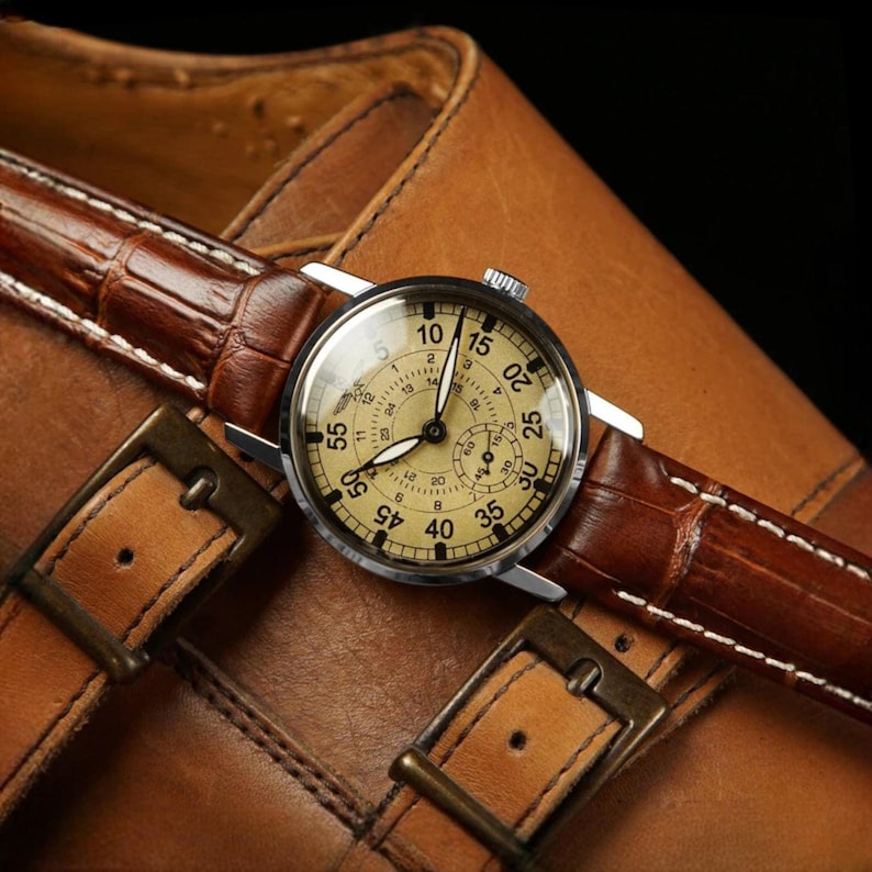 Puede incluir: Un reloj de pulsera vintage con esfera clara y n&uacute;meros negros, con una correa de cuero marr&oacute;n con estampado de cocodrilo. El reloj se encuentra sobre un zapato de cuero marr&oacute;n, con hebillas met&aacute;licas.