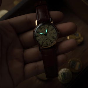 May include: A wristwatch with a round cream-coloured dial, black Arabic numerals, and a brown leather strap. The watch features luminous hour and minute hands that glow with a bright cyan light in the dark, set against a dark, low-light environment.