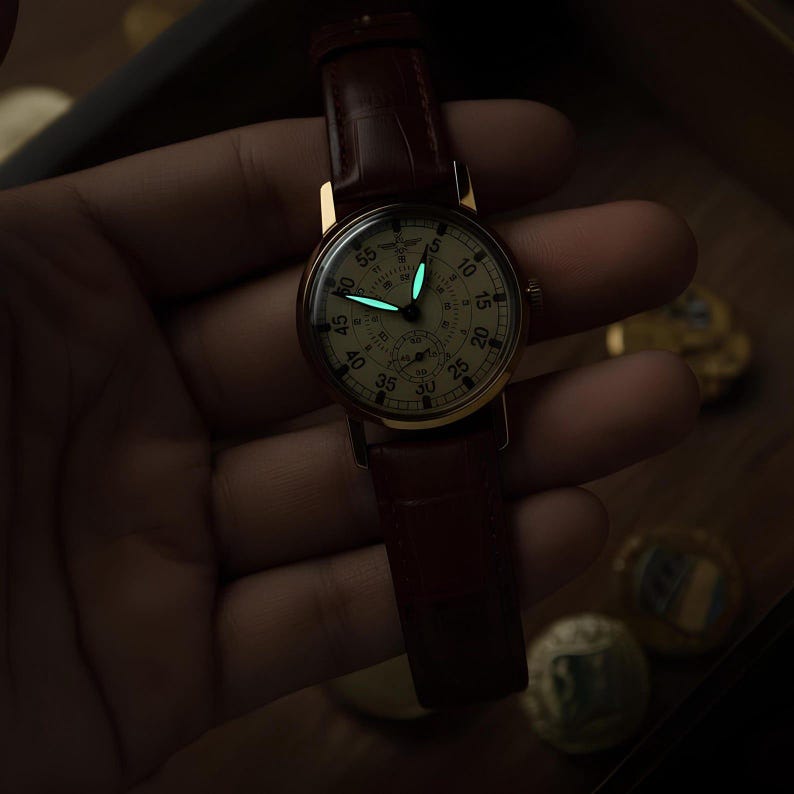 May include: A wristwatch with a round cream-coloured dial, black Arabic numerals, and a brown leather strap. The watch features luminous hour and minute hands that glow with a bright cyan light in the dark, set against a dark, low-light environment.