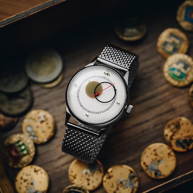 May include: A silver-tone wristwatch with a textured white dial, featuring a prominent gold circular element, a thin black ring, and a slender red second hand. The watch has a metallic mesh strap and a polished silver case with a side crown.