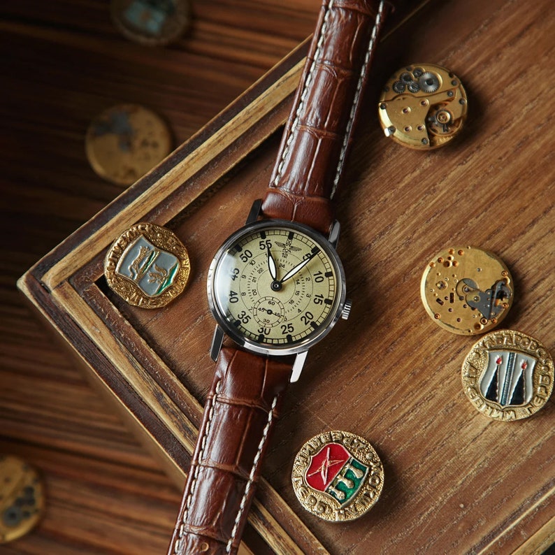May include: A vintage wristwatch with a brown leather band and a cream-colored face. The watch has black Roman numeral hour markers and a black second hand. The watch is on a wooden surface with other decorative items.