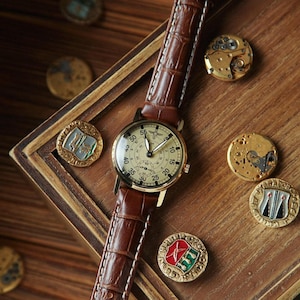 Puede incluir: Un reloj de pulsera de estilo vintage con una correa de cuero marr&oacute;n y una esfera crema. El reloj est&aacute; sobre una superficie de madera, rodeado de piezas de reloj antiguas y medallones decorativos. La esfera muestra n&uacute;meros y subesferas.