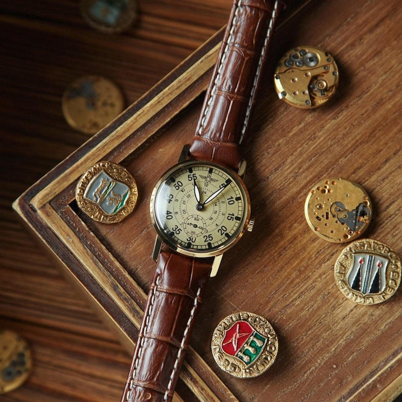 Puede incluir: Un reloj de pulsera de estilo vintage con una correa de cuero marr&oacute;n y una esfera crema. El reloj est&aacute; sobre una superficie de madera, rodeado de piezas de reloj antiguas y medallones decorativos. La esfera muestra n&uacute;meros y subesferas.