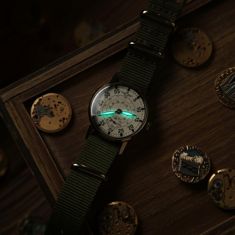 May include: A vintage-style wristwatch with a white face, black numerals, and a green nylon strap. The watch hands glow green in the dark. The watch is set against a dark wooden background with watch parts.