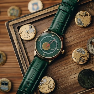 May include: A gold-toned wristwatch with a green face and a green leather band. The watch has a gold case and a red second hand. The watch is displayed on a wooden surface with watch components and decorative coins.
