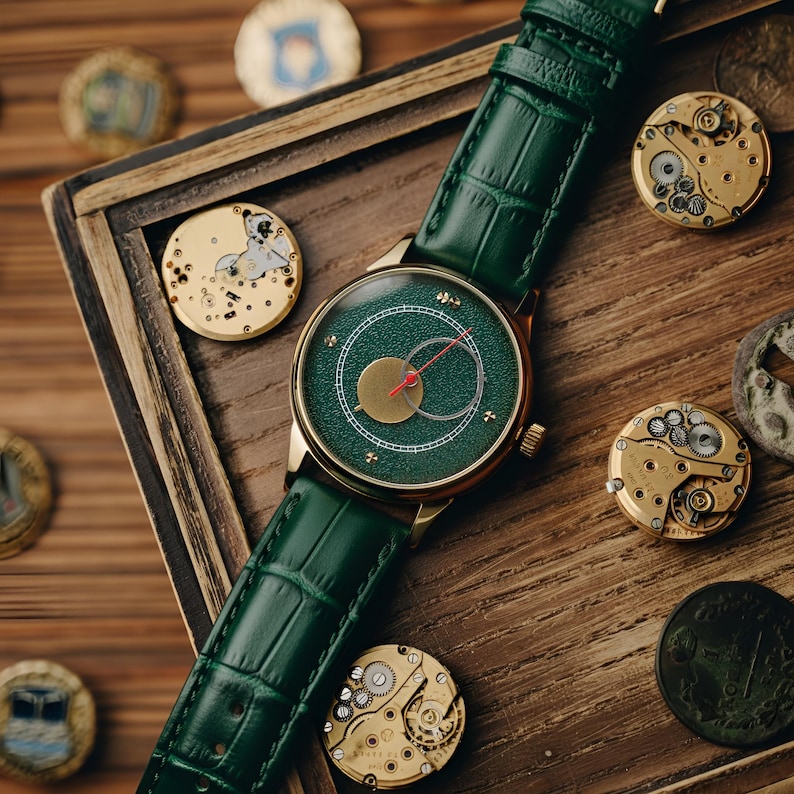 May include: A gold-toned wristwatch with a green face and a green leather band. The watch has a gold case and a red second hand. The watch is displayed on a wooden surface with watch components and decorative coins.