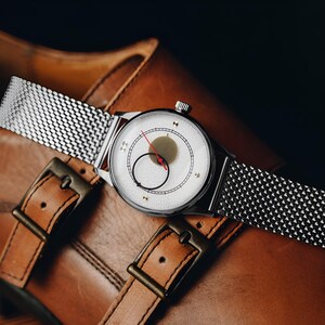 May include: A wristwatch with a silver-toned mesh strap and a round white textured dial. The dial features a central gold-toned circle, a black circular outline, and a thin red second hand. The watch case is polished silver-toned metal.