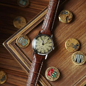 Puede incluir: Un reloj de pulsera de estilo vintage con una correa de cuero marr&oacute;n y una esfera crema. El reloj tiene una caja negra y un segundero. Se muestra sobre una superficie de madera con varios medallones antiguos.