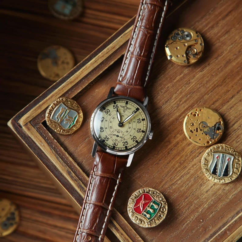 Puede incluir: Un reloj de pulsera de estilo vintage con una correa de cuero marr&oacute;n y una esfera crema. El reloj tiene una caja negra y un segundero. Se muestra sobre una superficie de madera con varios medallones antiguos.