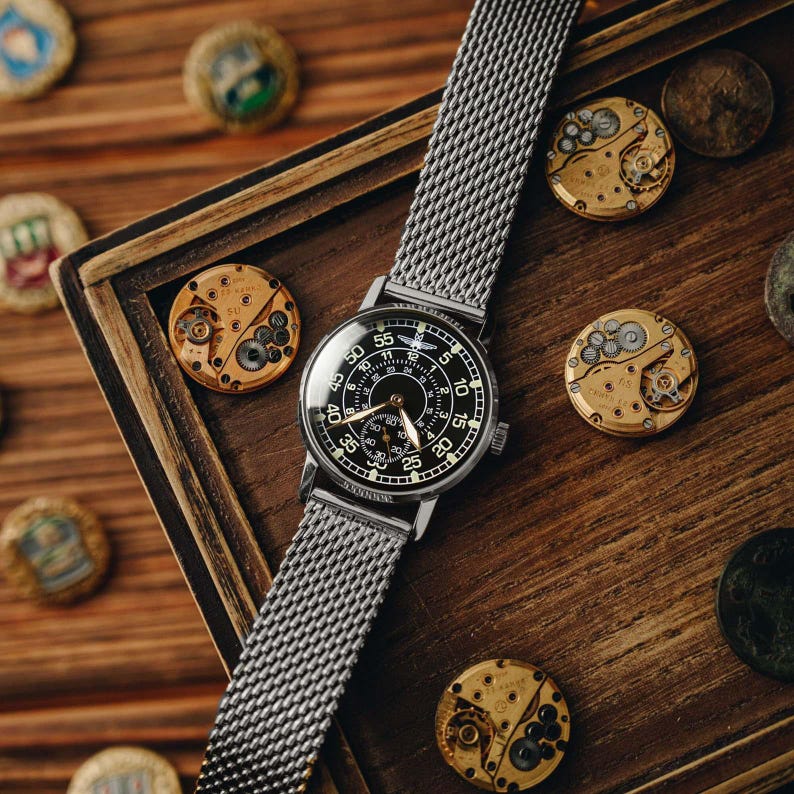 May include: A silver wristwatch with a black face and mesh band rests on a wooden surface. The watch face displays white numerals and hands. Several watch movement components and bottle caps are scattered around the watch.