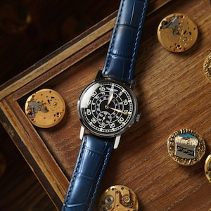 Puede incluir: Un reloj de pulsera con esfera negra, n&uacute;meros blancos y una subesfera, con una correa de cuero azul. El reloj se exhibe sobre una superficie de madera, con piezas de reloj y alfileres decorativos esparcidos alrededor.
