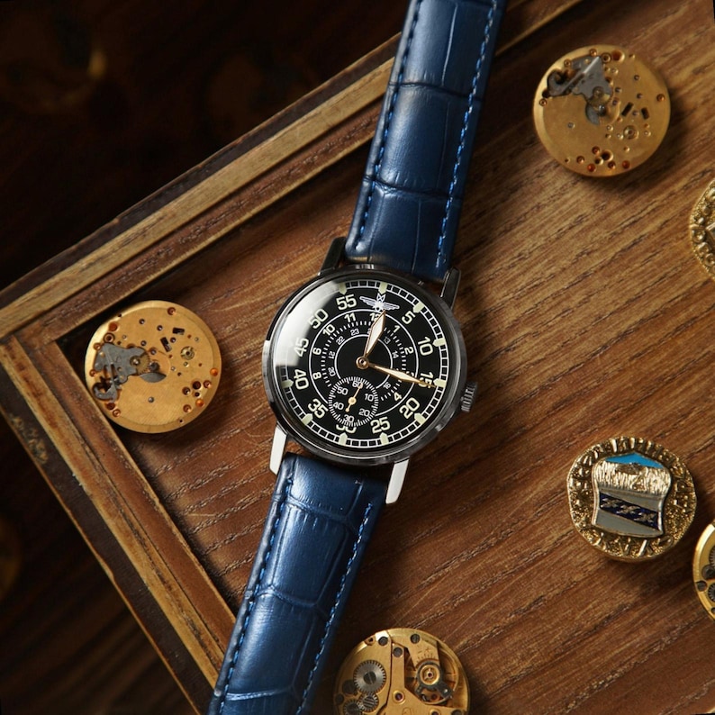 Puede incluir: Un reloj de pulsera con esfera negra, n&uacute;meros blancos y una subesfera, con una correa de cuero azul. El reloj se exhibe sobre una superficie de madera, con piezas de reloj y alfileres decorativos esparcidos alrededor.