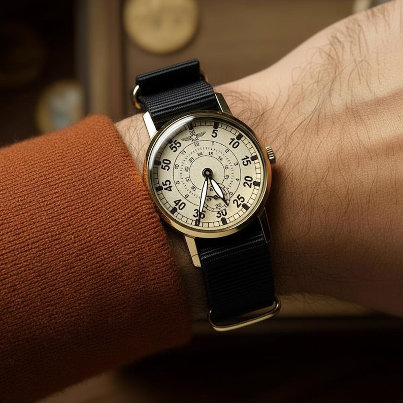 May include: A gold-toned wristwatch with a black nylon strap. The watch face is cream-colored with black numerals and hands. The watch is on a person's wrist, showing the time. The watch has a classic, vintage aesthetic.