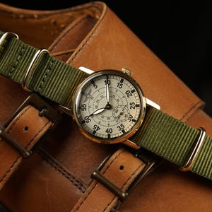 May include: A gold-toned wristwatch with a cream-colored face and black numerals, featuring a green nylon strap. The watch is set against a backdrop of brown leather, showcasing the details of the watch and the strap.