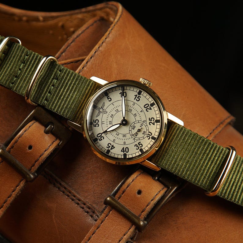 May include: A gold-toned wristwatch with a cream-colored face and black numerals, featuring a green nylon strap. The watch is set against a backdrop of brown leather, showcasing the details of the watch and the strap.