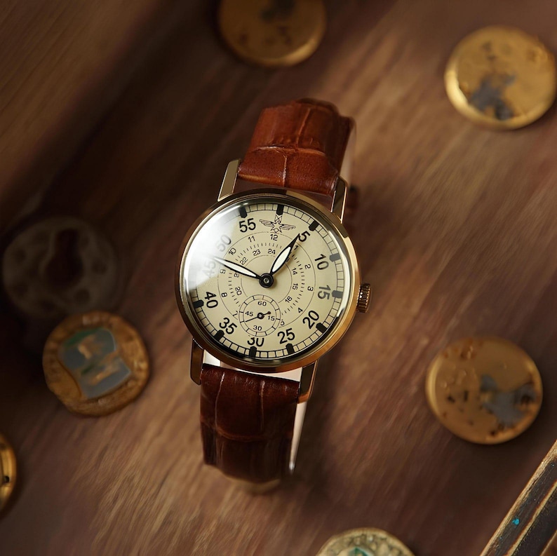 May include: A wristwatch with a round gold-tone case and a cream-coloured dial featuring black numerals, a small sub-dial, and a winged logo. The watch is fitted with a brown leather strap that has a textured, embossed pattern on its surface.