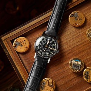 May include: A black wristwatch with a black leather band, featuring a black dial with white numerals and hands. The watch is displayed on a wooden surface with several gold-colored watch components.