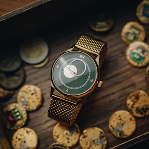 May include: A gold-toned wristwatch with a green face and a mesh band. The watch face features a white crescent design and small cross-shaped markers. The watch is surrounded by gold-colored coins on a wooden surface.
