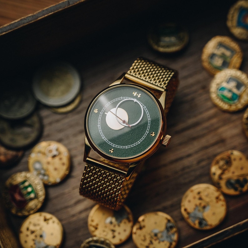 May include: A gold-toned wristwatch with a green face and a mesh band. The watch face features a white crescent design and small cross-shaped markers. The watch is surrounded by gold-colored coins on a wooden surface.